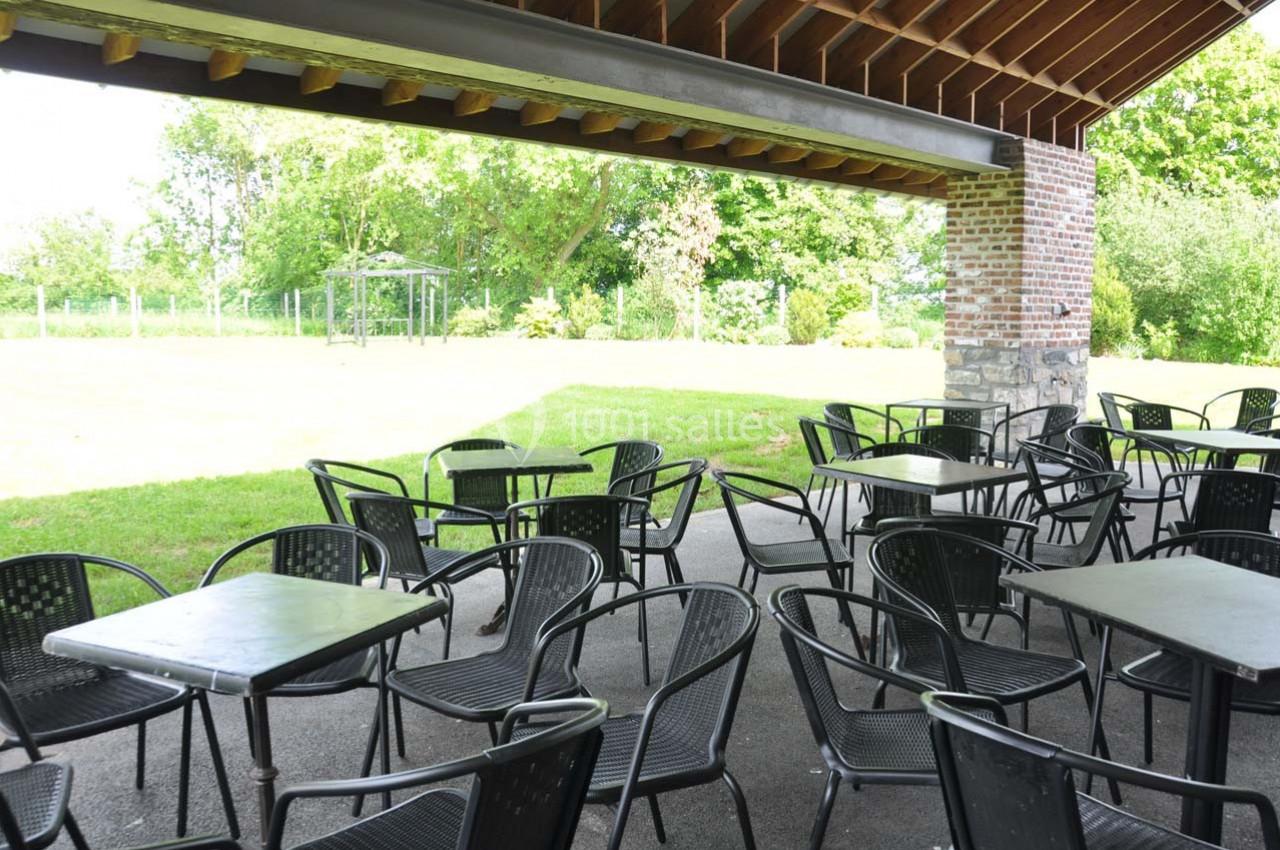 Terrasse couverte avec des tables et chaises noires, donnant sur un jardin verdoyant par une journée ensoleillée.
