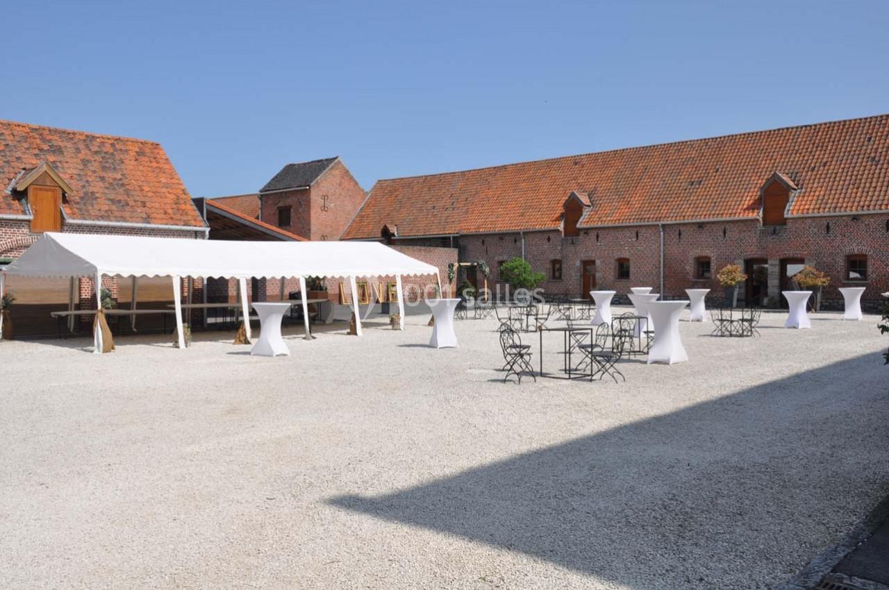 Cour intérieure d'une ferme rénovée avec tables hautes, chaises en fer forgé et une tente blanche sous un ciel dégagé.