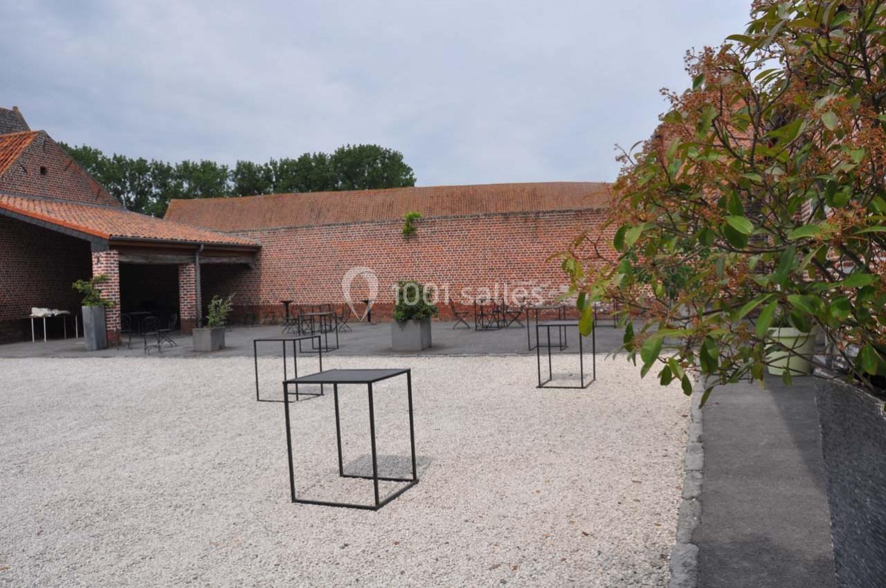 Cour extérieure avec gravier, tables hautes noires, murs en briques rouges et végétation en arrière-plan.