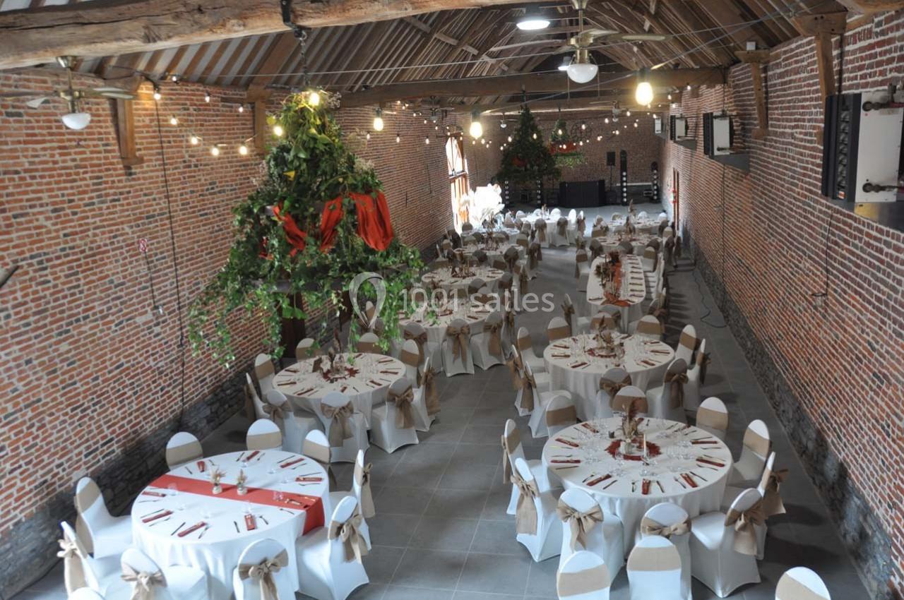 Salle de réception en briques avec tables rondes décorées, chaises blanches et guirlandes lumineuses suspendues.