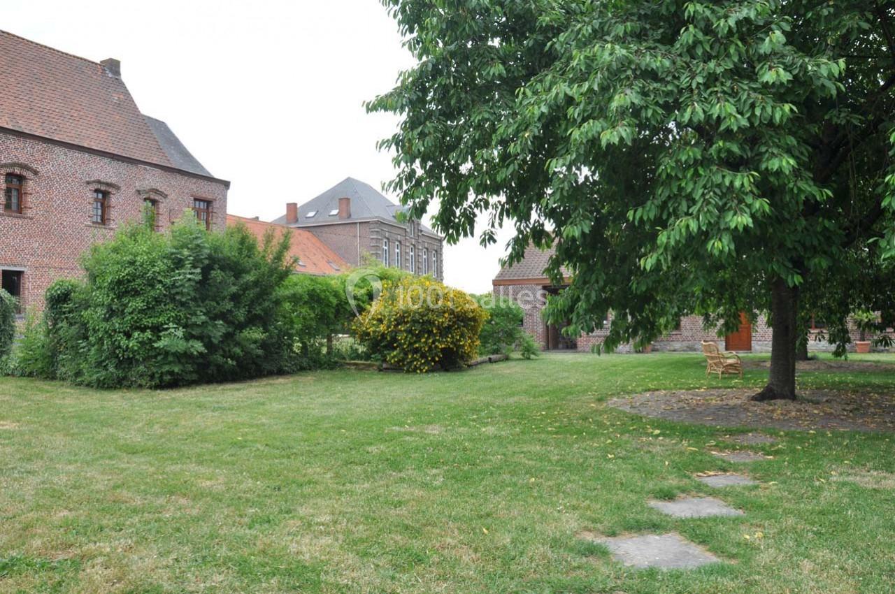 Jardin verdoyant avec pelouse, arbres, arbustes et bâtiments en briques rouges en arrière-plan.