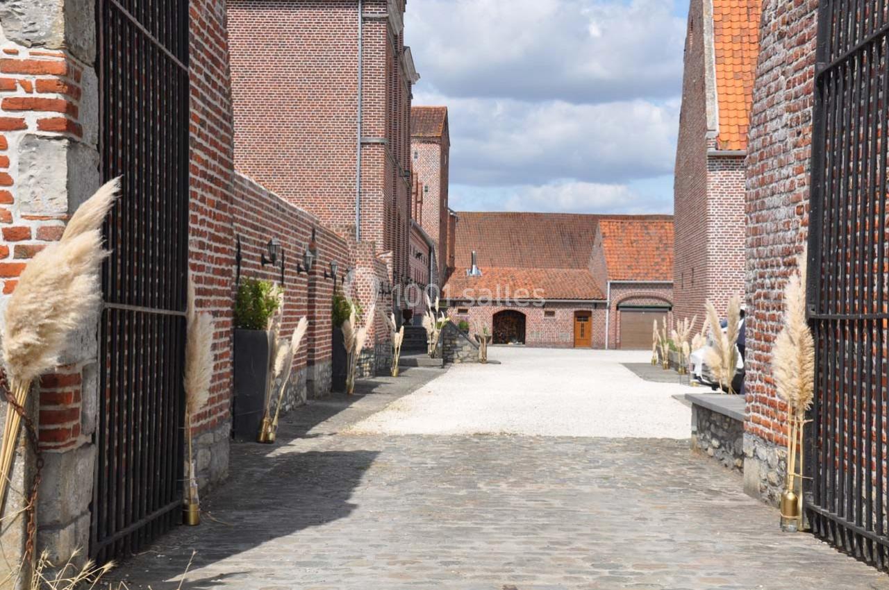 Allée pavée bordée de bâtiments en briques rouges et de graminées décoratives, sous un ciel partiellement nuageux.
