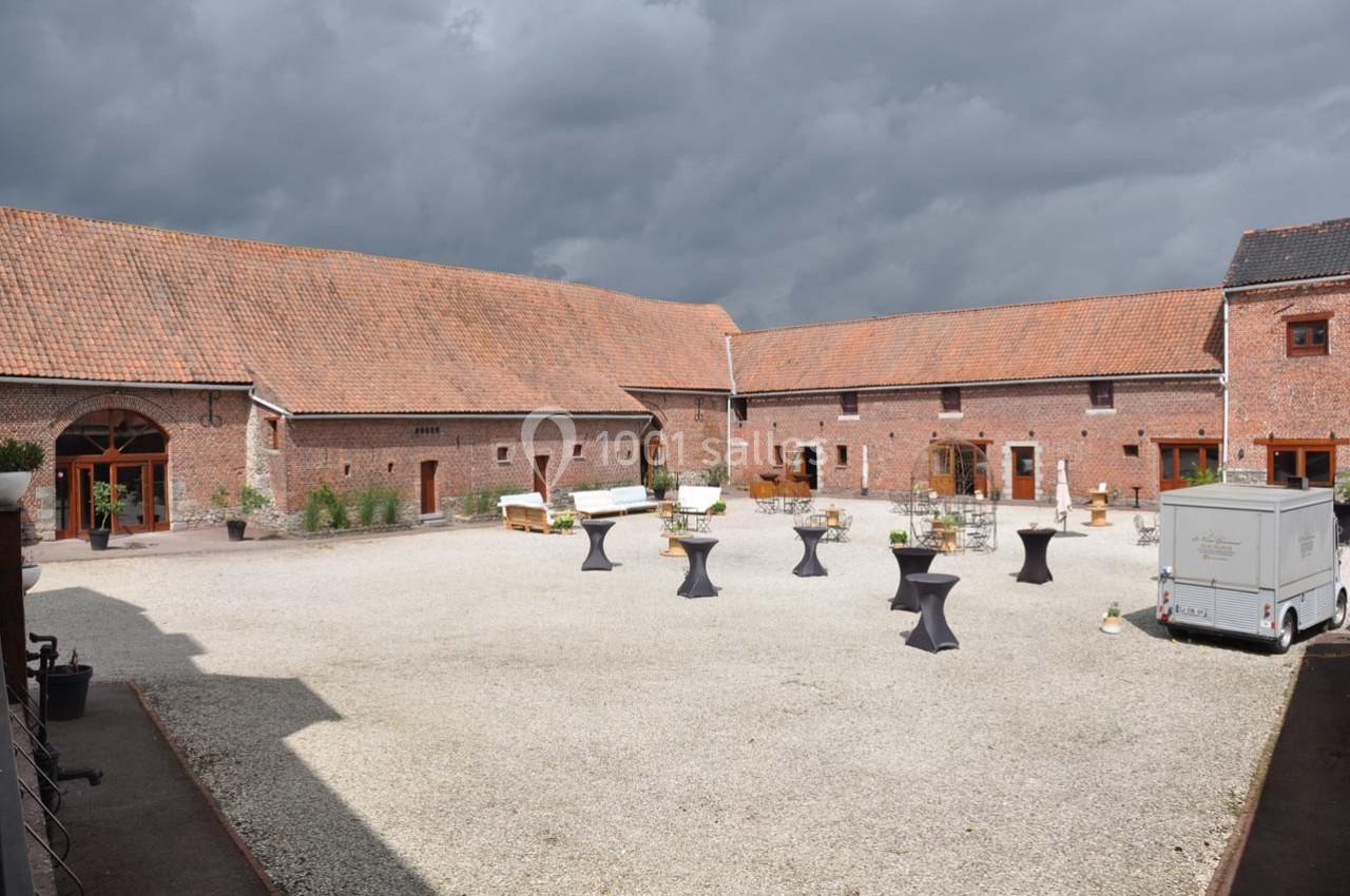 Cour intérieure d'une ferme rénovée avec pavés, tables hautes noires et un ciel nuageux en arrière-plan.