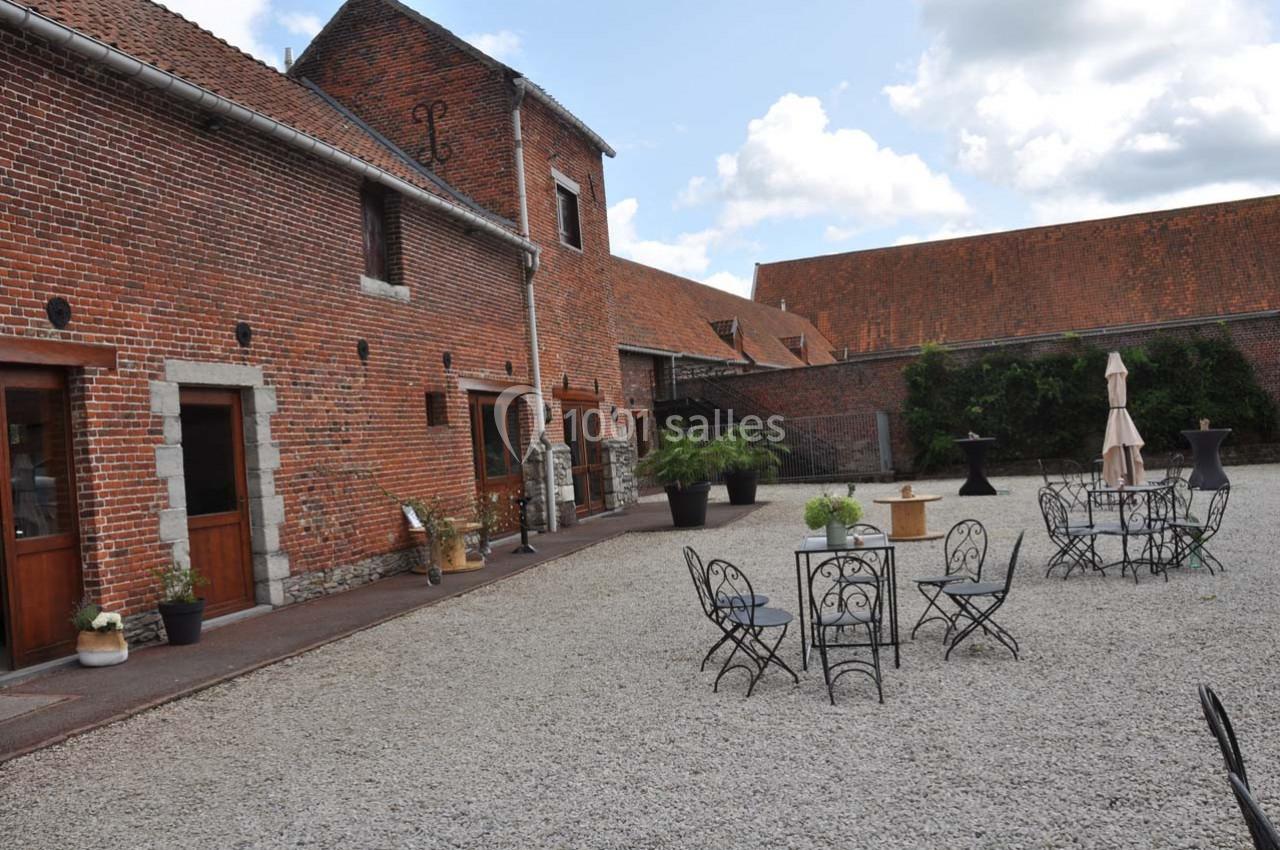 Cour pavée avec mobilier de jardin en fer forgé, entourée de bâtiments en briques rouges sous un ciel partiellement nuageux.
