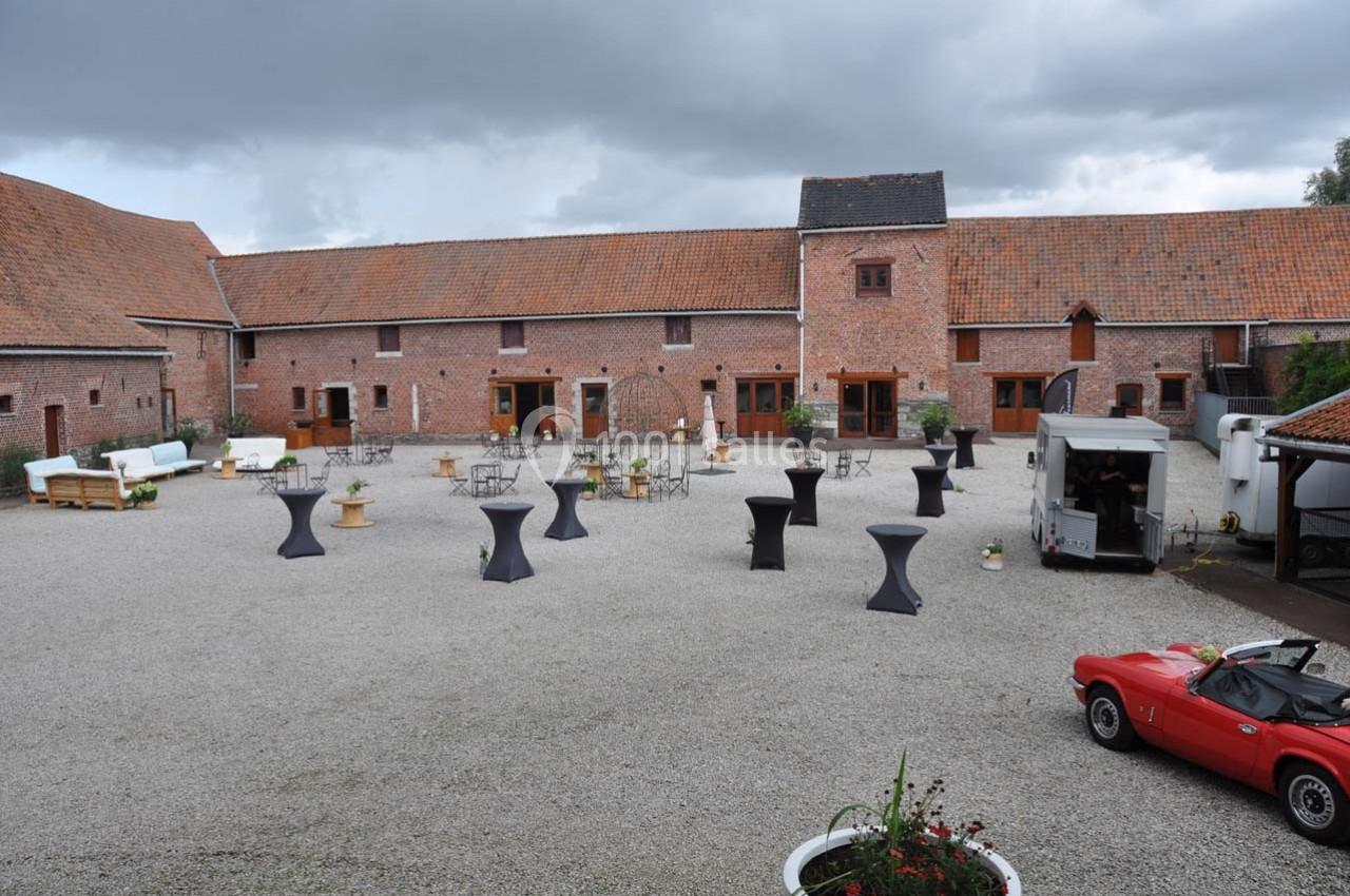 Cour intérieure d'une ferme rénovée avec tables hautes, mobilier de réception et une voiture ancienne rouge.