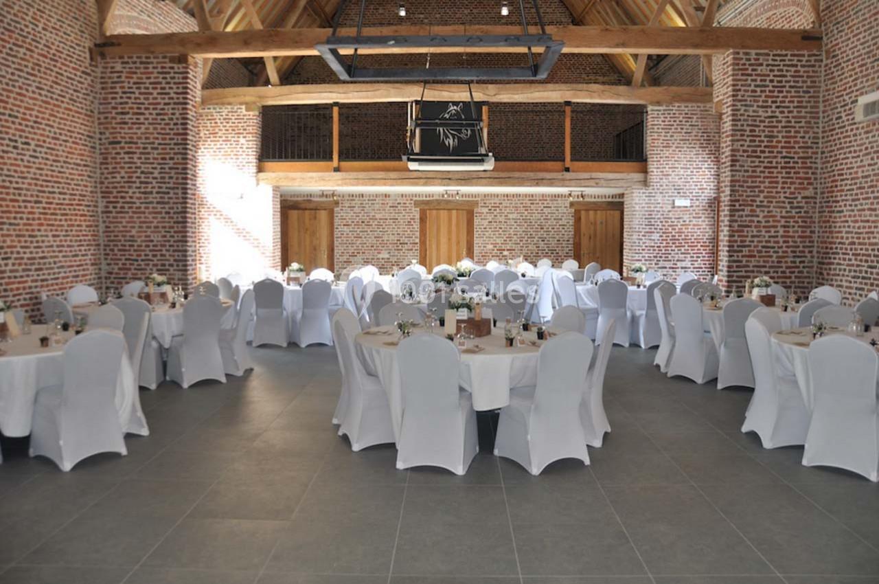 Salle de réception avec murs en briques, tables rondes dressées et chaises blanches, éclairée par la lumière naturelle.