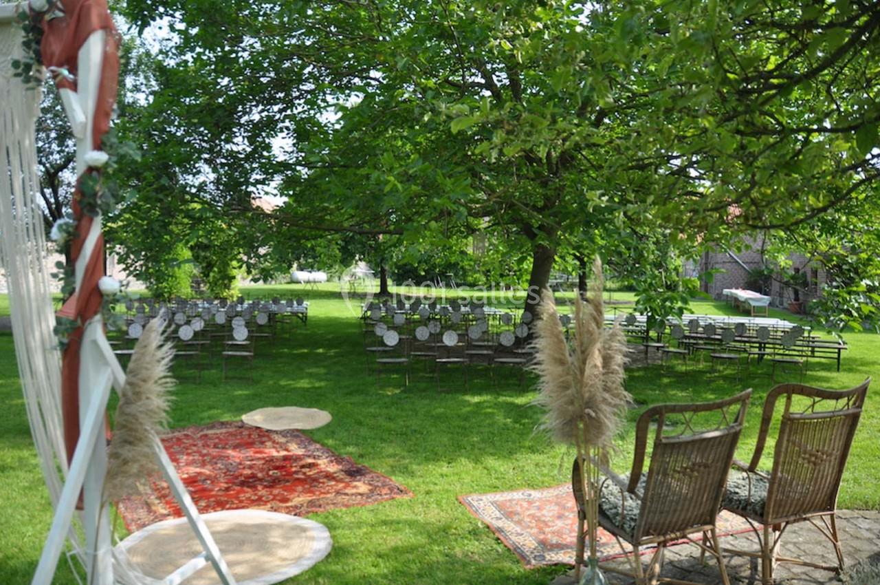 Chaises et tables disposées en extérieur sous des arbres, décorées pour un événement sur une pelouse avec tapis et fauteuils.