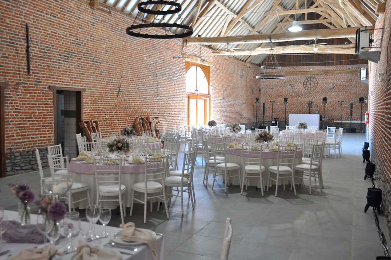 Salle de réception en briques avec tables rondes décorées, chaises blanches et poutres apparentes au plafond.