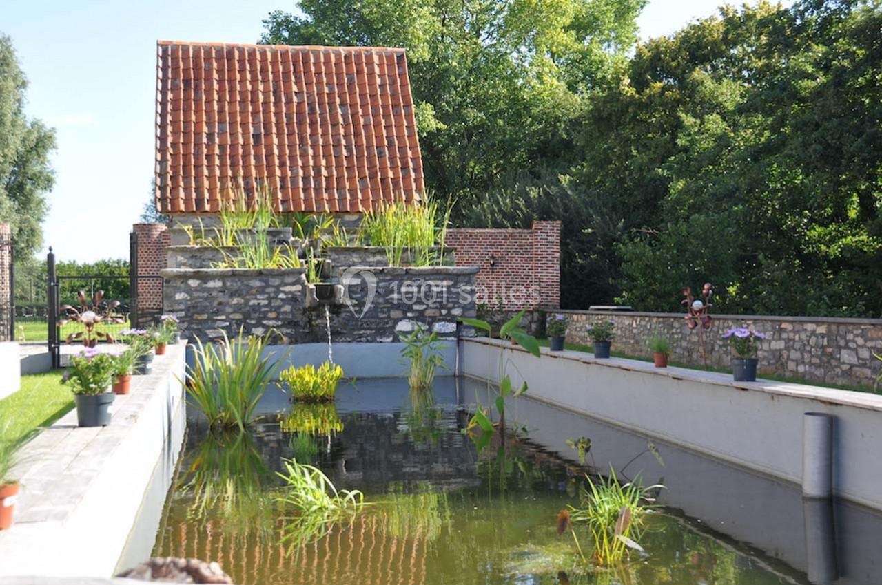 Bassin rectangulaire avec plantes aquatiques, bordé de murs en pierre et d'un bâtiment en briques au toit rouge.