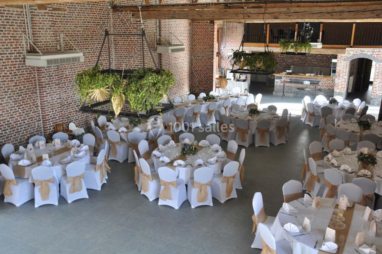 Salle de réception décorée avec des tables rondes dressées, chaises blanches avec nœuds dorés et plantes suspendues.