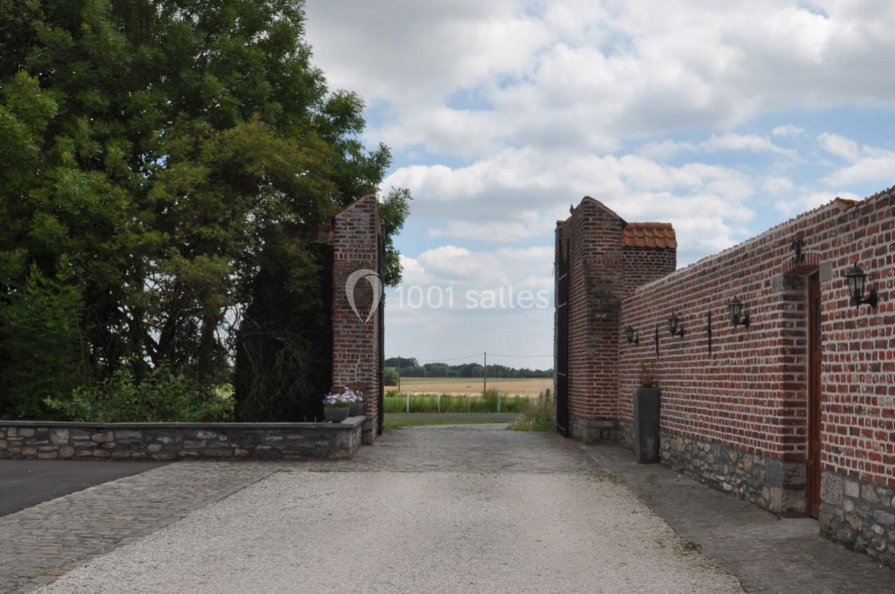 Entrée d'une cour en briques rouges avec un chemin en gravier, bordée d'arbres et donnant sur un paysage champêtre.