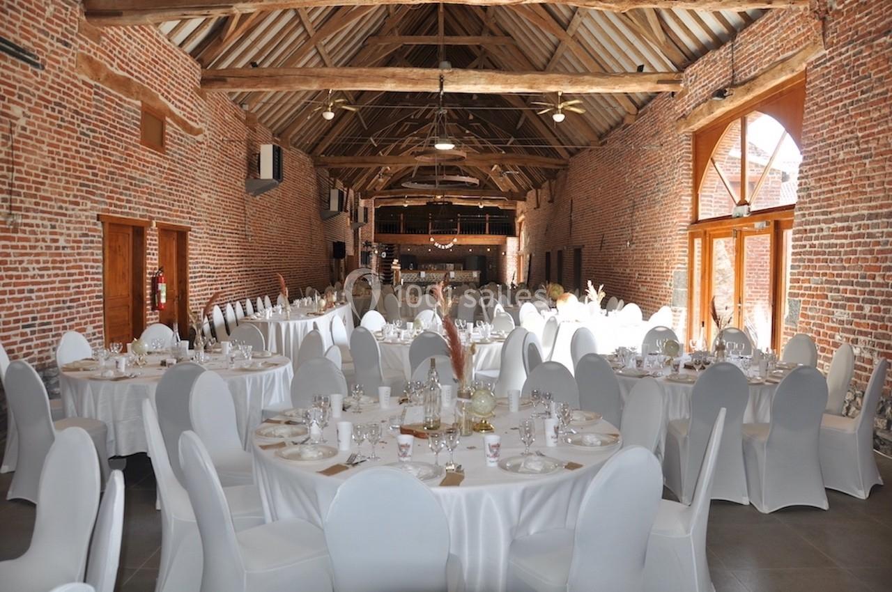 Salle de réception en briques avec tables rondes dressées, nappes blanches et chaises couvertes, sous une charpente en bois.