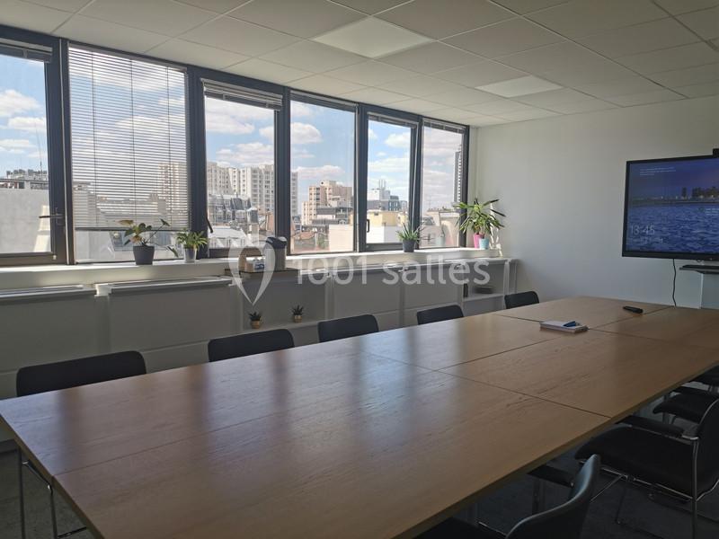 Salle de réunion lumineuse avec une grande table en bois, des chaises noires, des plantes et une vue sur des immeubles…
