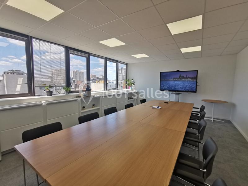 Salle de réunion lumineuse avec une grande table en bois, des chaises noires, un écran et des fenêtres donnant sur la ville.