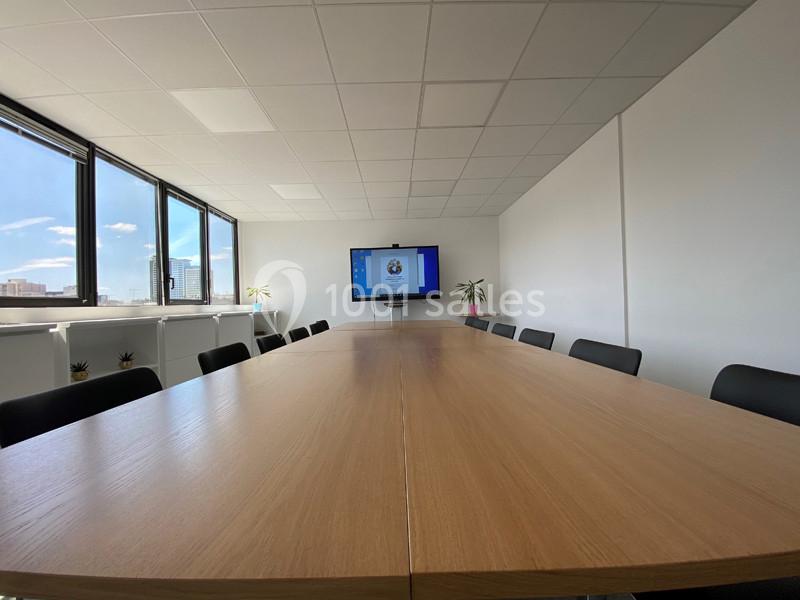 Salle de réunion lumineuse avec une grande table en bois, des chaises noires et un écran affichant une présentation.