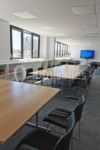 Salle de réunion lumineuse avec grandes tables en bois, chaises noires, étagères blanches et écran mural.