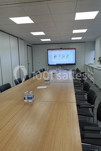 Salle de réunion avec une longue table en bois, des chaises noires, des bouteilles d'eau et un écran affichant une…