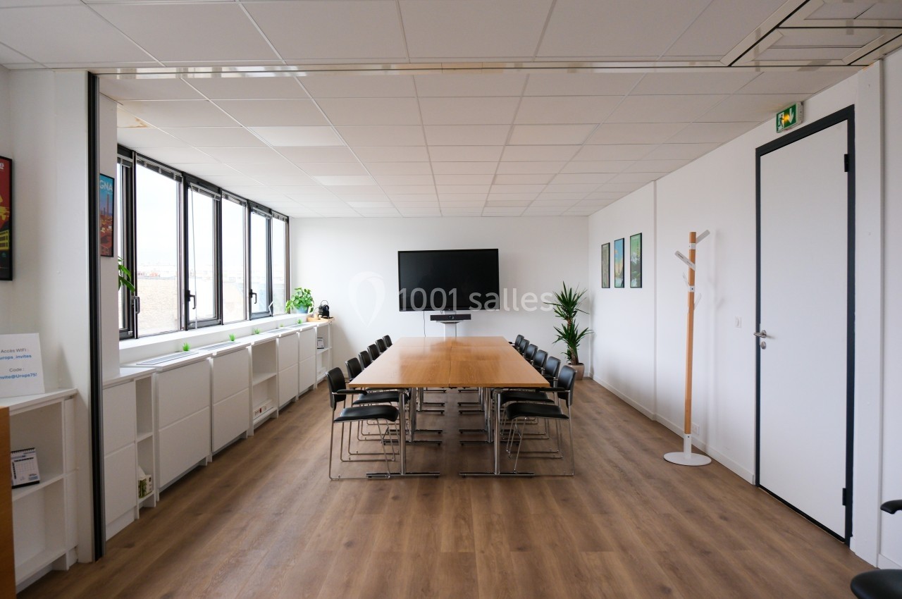 Salle de réunion lumineuse avec une grande table en bois, des chaises noires, un écran mural et des plantes décoratives.