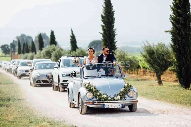 Un couple de mariés souriants dans une voiture décapotable décorée, suivi d'un cortège sur une route de campagne.