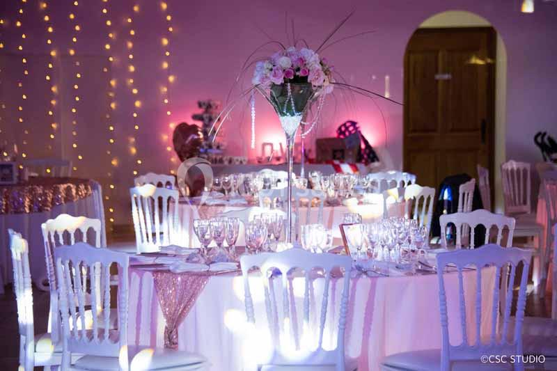 Salle de réception décorée avec des tables rondes, nappes blanches, chaises élégantes et éclairage rose tamisé.