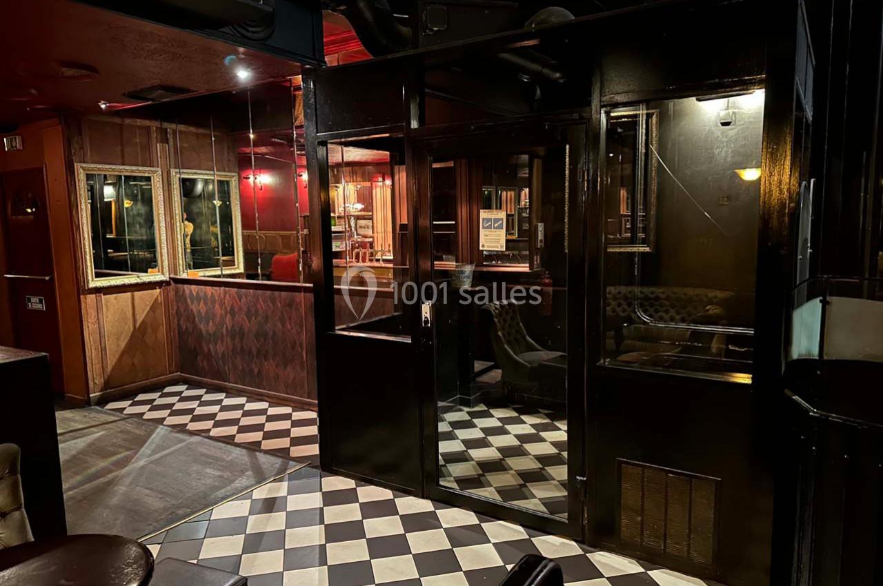 Intérieur d'un bar avec sol en damier, murs sombres, éclairage tamisé et mobilier en bois et cuir.