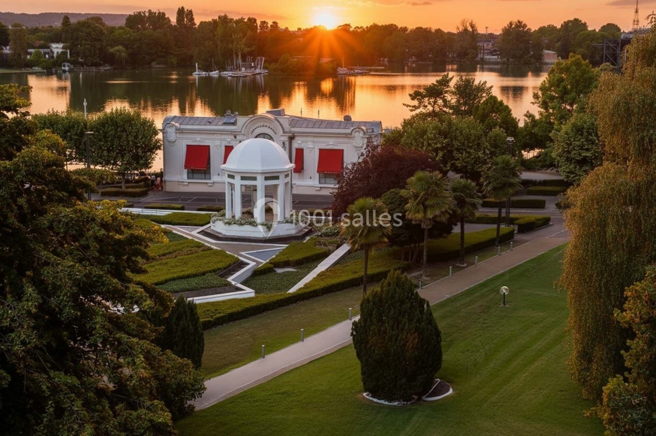 Vue d'un parc paysager avec un kiosque blanc, des allées bordées d'arbres et un lac au coucher du soleil.