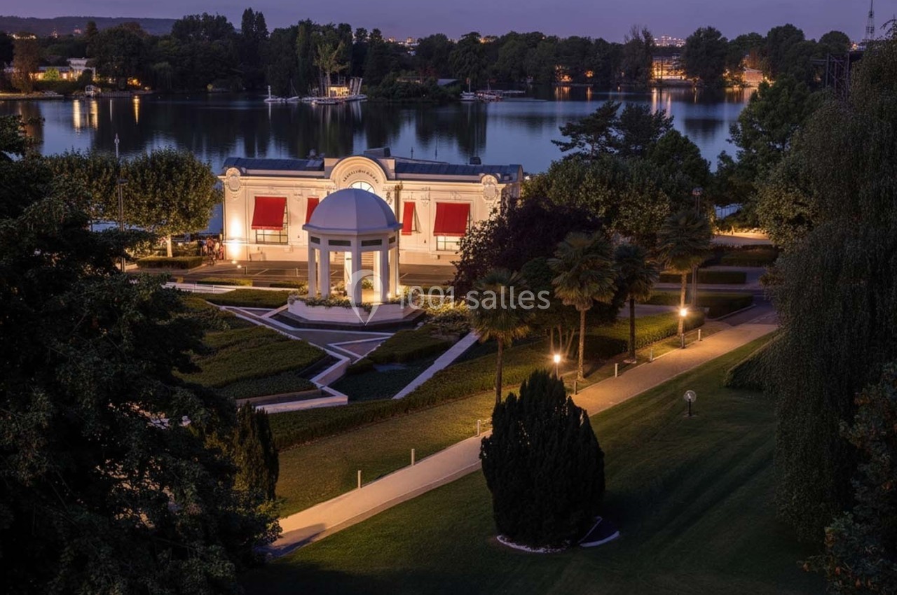 Vue nocturne d'un parc avec un pavillon éclairé, des allées bordées d'arbres et un plan d'eau en arrière-plan.
