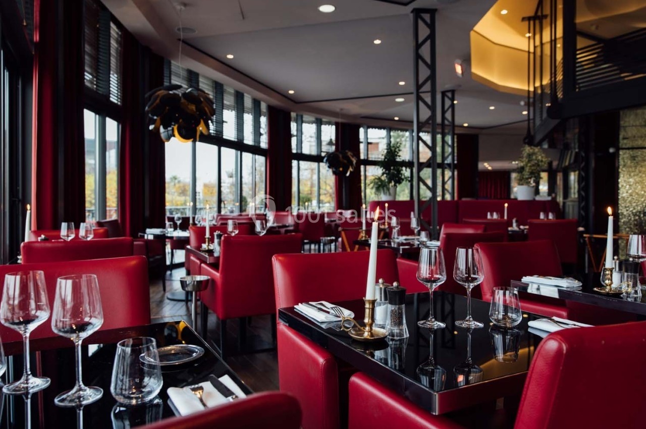 Salle de restaurant élégante avec des tables noires, des chaises rouges, des bougies et de grandes baies vitrées.