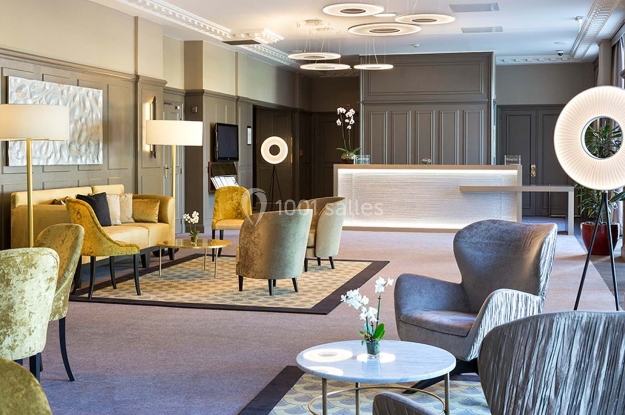 Salon d'hôtel élégant avec fauteuils, tables basses, éclairage moderne et comptoir de réception en arrière-plan.