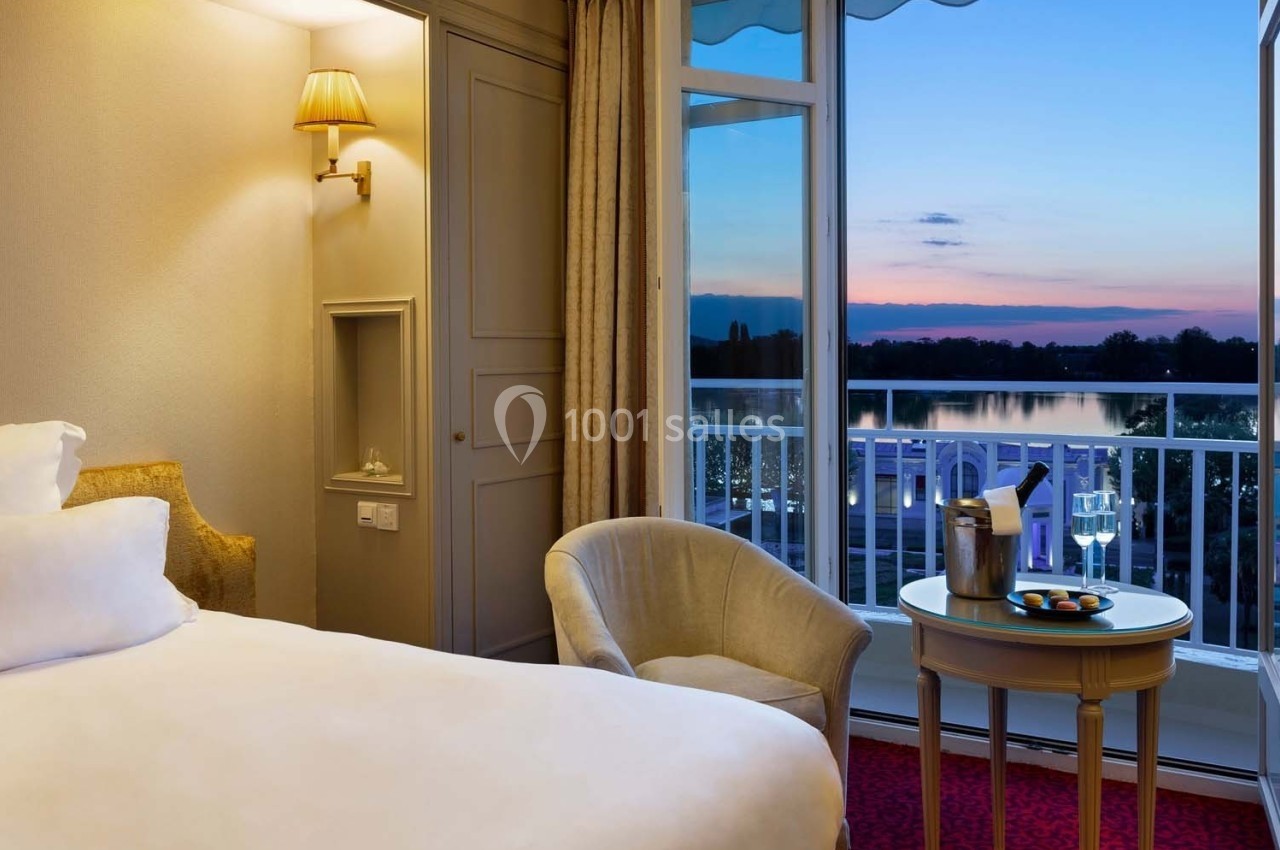 Chambre d'hôtel avec lit, fauteuil, table dressée et vue sur un balcon donnant sur un paysage au coucher du soleil.