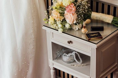 Robe de mariée, bouquet de fleurs, chaussures et bijoux posés près d'une table sur tapis rouge.