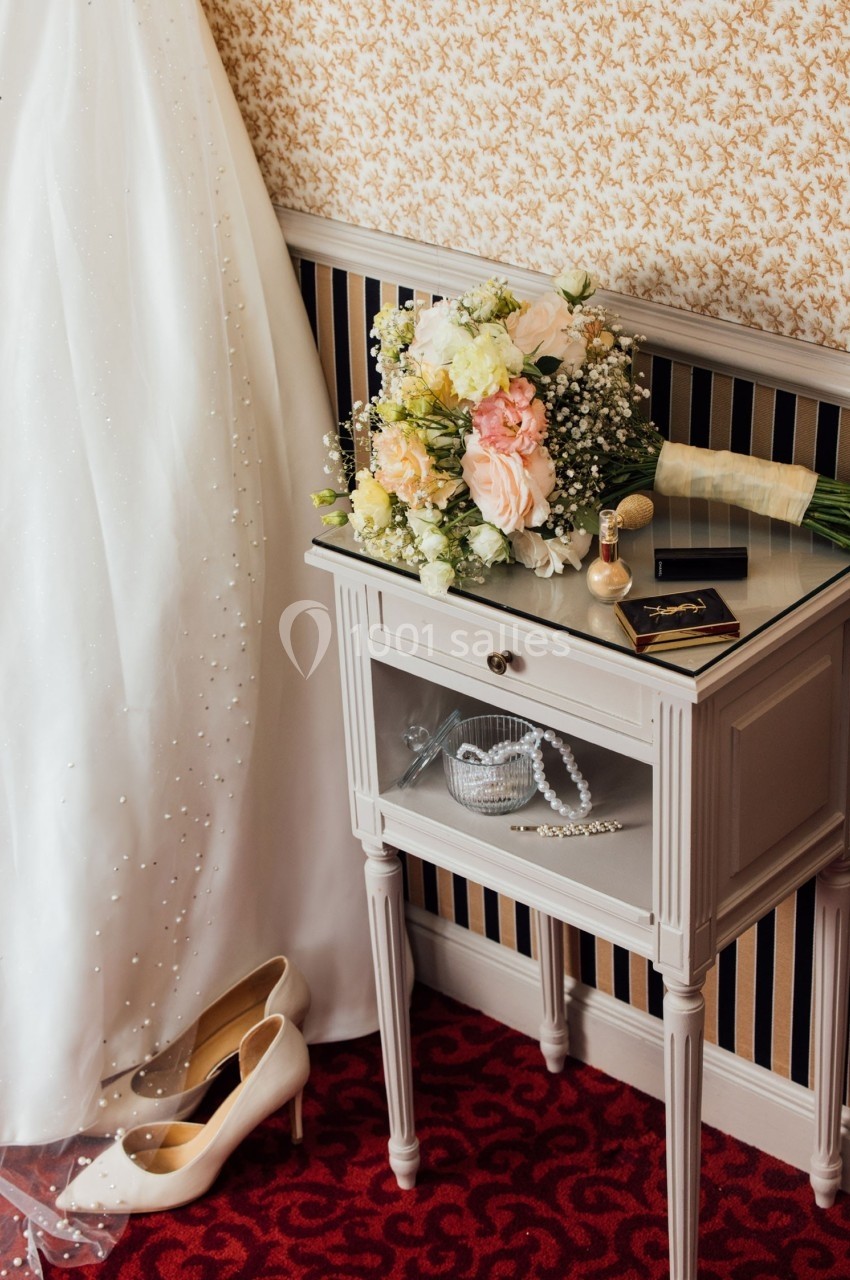 Robe de mariée, bouquet de fleurs, chaussures et bijoux posés près d'une table sur tapis rouge.