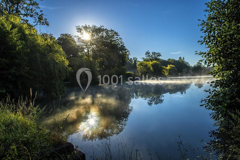 Vue d'un lac entouré d'arbres au lever du soleil, avec des reflets sur l'eau et une légère brume.