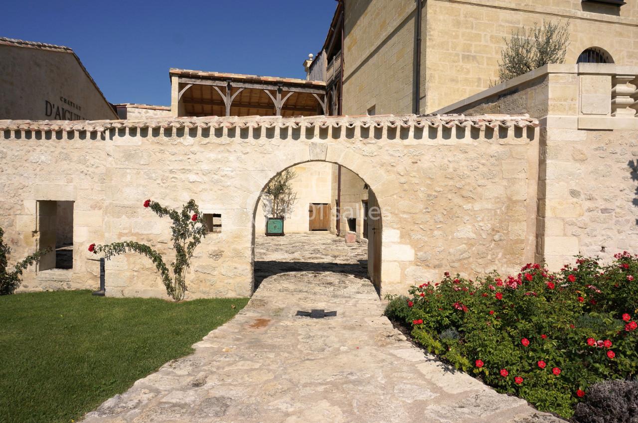 Cour en pierre avec arche, murs en pierre claire, pelouse, rosiers rouges et bâtiment en arrière-plan sous un ciel bleu.