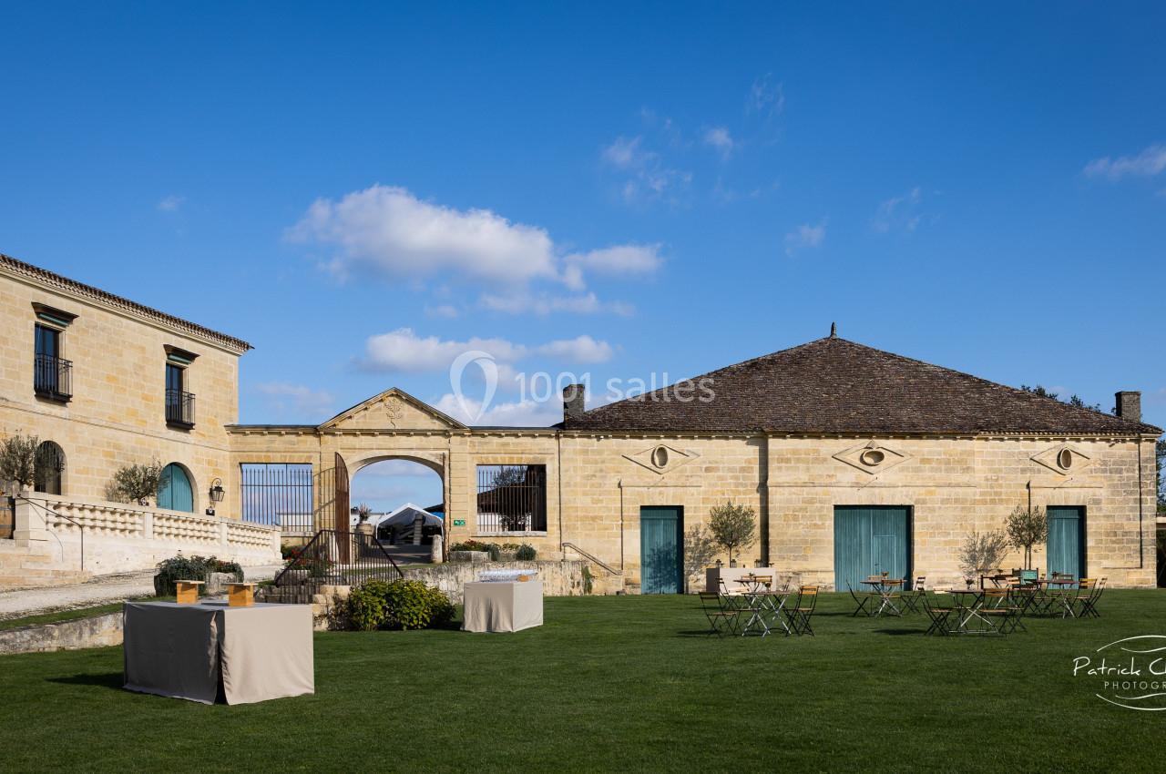 Cour d'un domaine en pierre avec bâtiments anciens, pelouse aménagée, tables et chaises, sous un ciel bleu.