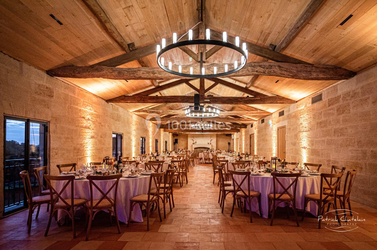 Salle de réception en pierre avec tables dressées, chaises en bois et lustres circulaires suspendus au plafond en poutres…