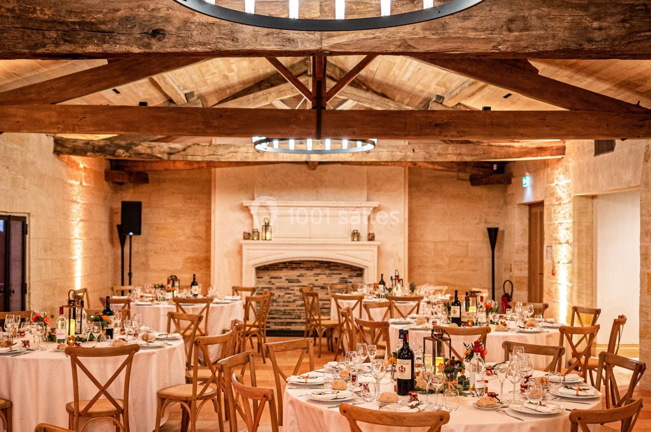 Salle de réception chaleureuse avec tables rondes dressées, chaises en bois et un lustre circulaire suspendu.