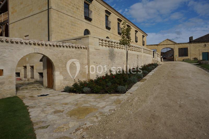 Allée en gravier bordée de fleurs et d'arbustes, longeant un bâtiment en pierre avec des arcades et fenêtres.