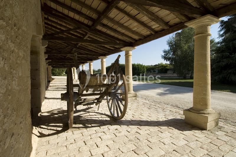Charrette en bois sous un auvent en pierre, sur un sol pavé, avec un jardin visible en arrière-plan.
