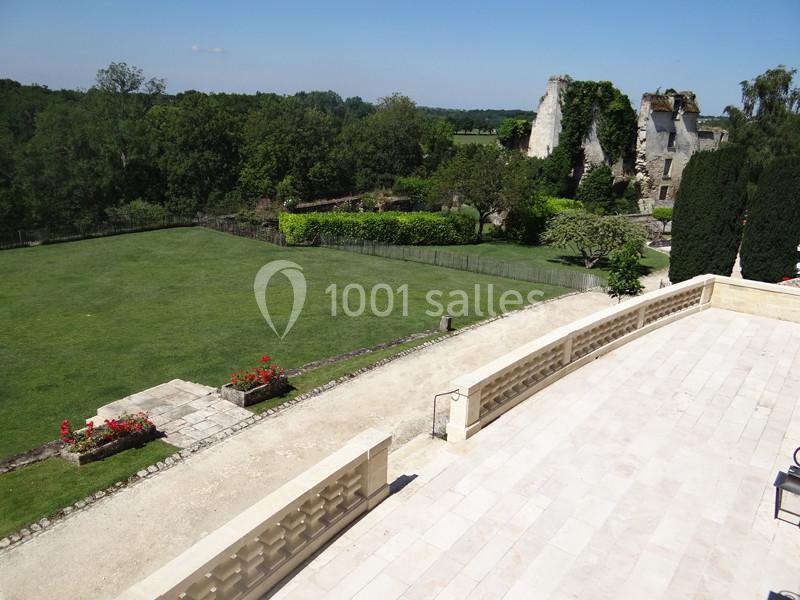 Vue d'une terrasse en pierre donnant sur un jardin verdoyant avec des ruines anciennes en arrière-plan.