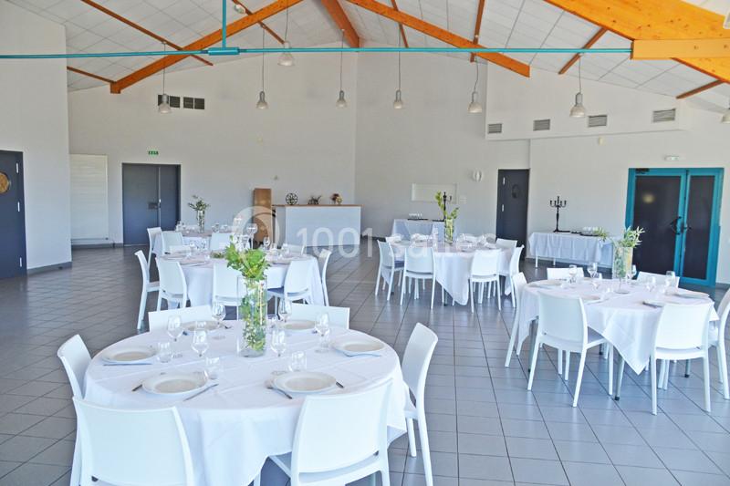 Salle lumineuse avec tables rondes dressées, chaises blanches, poutres apparentes et décoration florale simple.