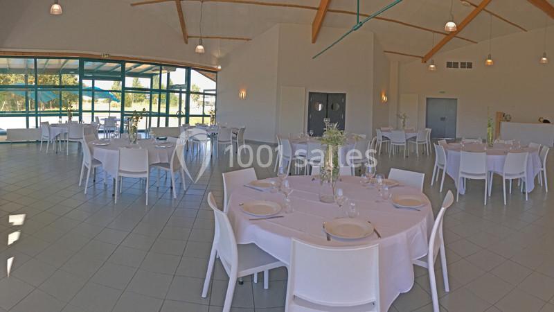 Salle de réception lumineuse avec tables rondes dressées, chaises blanches et grandes baies vitrées donnant sur l'extérieur.
