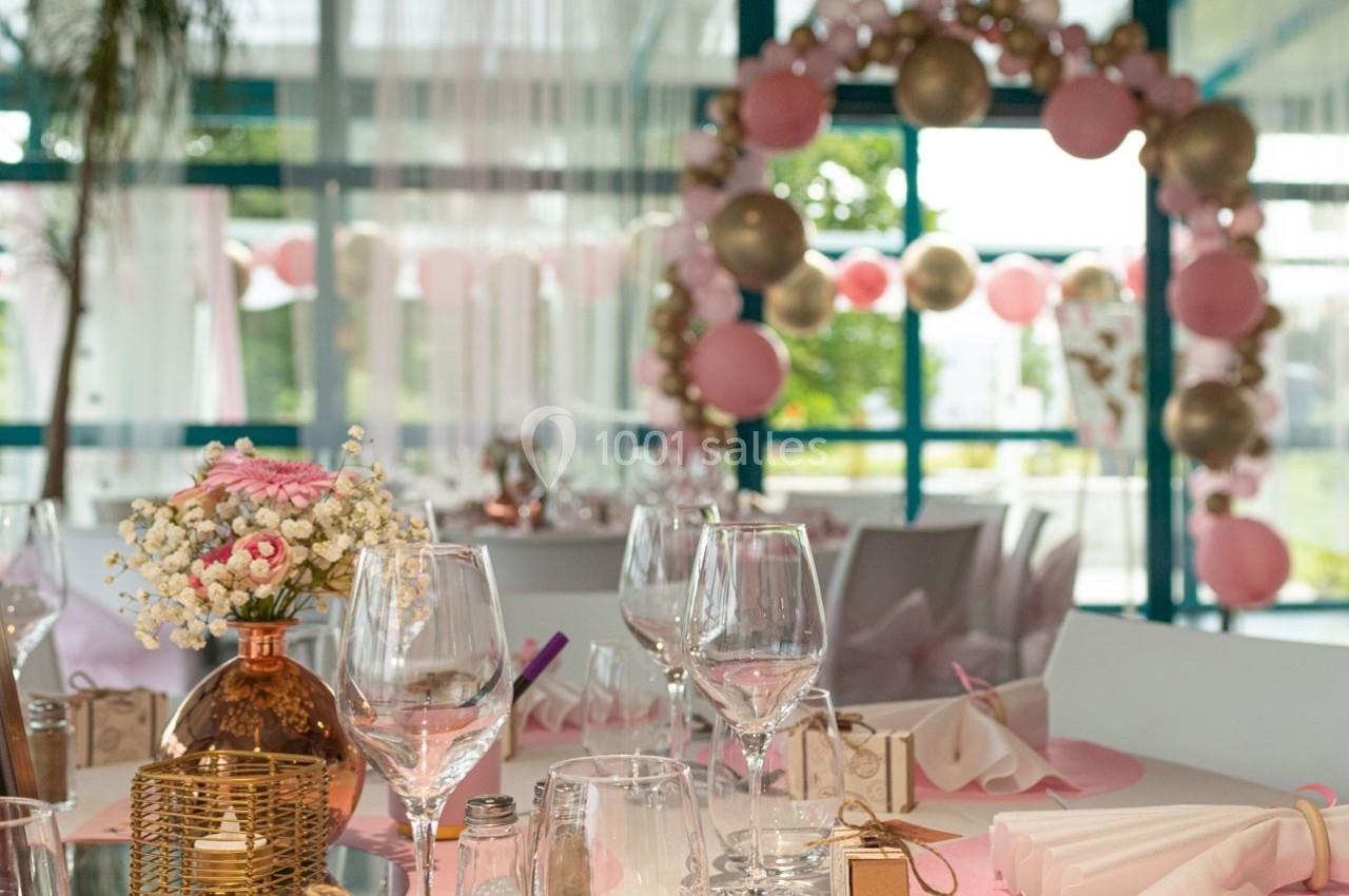 Table décorée avec des verres, des fleurs et des cadeaux, devant une arche de ballons roses et dorés dans une salle…