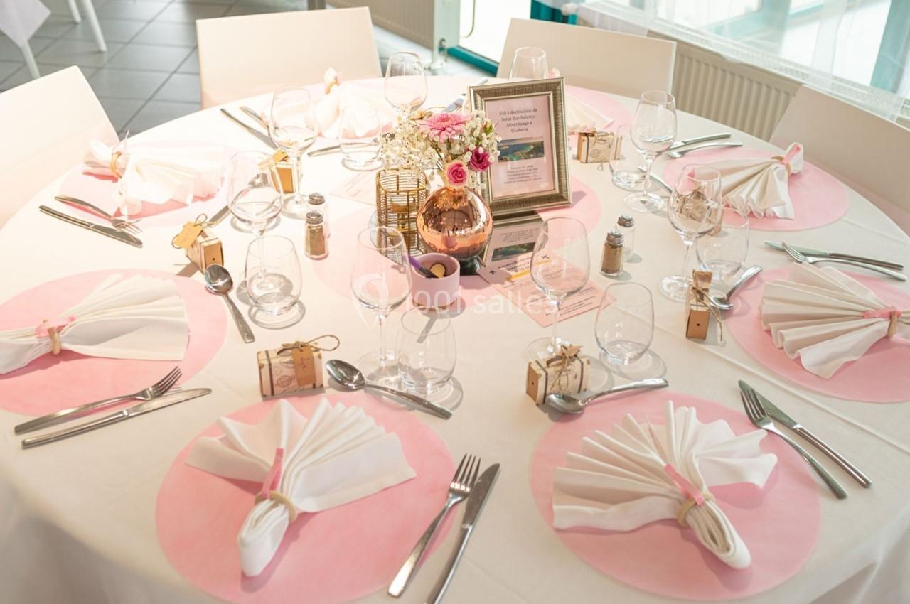 Table décorée pour un événement, avec nappes blanches, sets de table roses, serviettes pliées et centre de table floral.
