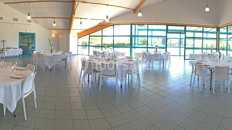 Salle lumineuse avec de grandes baies vitrées, tables rondes dressées avec nappes blanches et chaises modernes.
