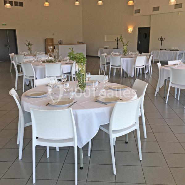 Salle de réception lumineuse avec des tables rondes dressées de nappes blanches, vaisselle et décorations simples.