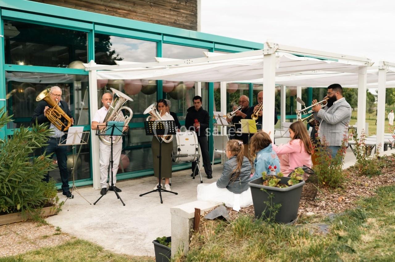 Un groupe de musiciens joue en plein air sous une pergola, devant un bâtiment vitré, avec des spectateurs assis.