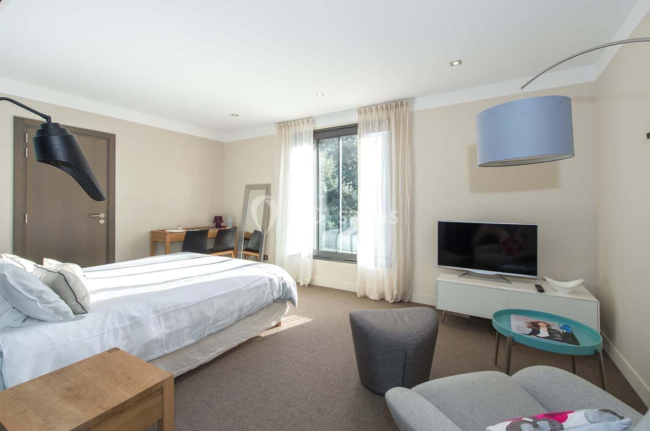 Chambre lumineuse avec lit double, bureau, télévision, fauteuil et grande fenêtre avec rideaux clairs.