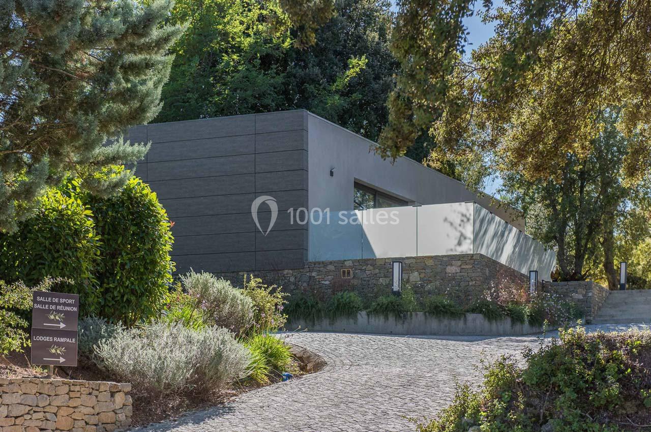 Bâtiment moderne entouré de végétation, avec un chemin pavé et des panneaux indiquant des salles et des logements.