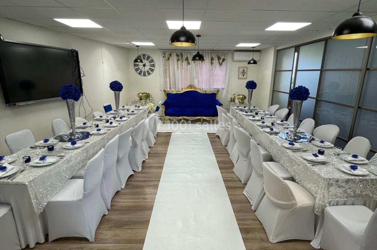 Salle décorée pour un événement, avec tables alignées, nappes blanches, chaises houssées et accents bleus.
